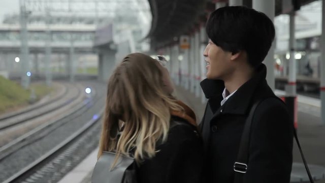 The multiathnical couple is hugging and enjoying the meeting in the railway station. The young korean man in black coat is tlking to his european girlfriend and cuddling her. The woman is wearing sand