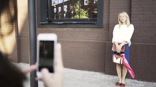 Unrecognizable dark haired woman making pictures of a blonde model wearing a short skirt and a blouse and smiling in a city street. Right to left pan real time medium shot