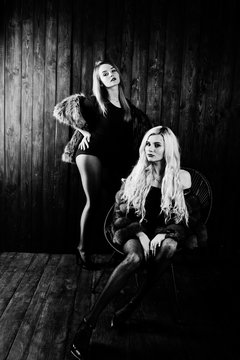 Two elegante sexy blonde girls wear on fur coat sitting on chair at studio against wooden background.