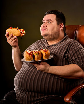 Fat man eating fast food hot dog on plate. Breakfast for overweight person. Junk meal leads to obesity. Person regularly overeats concept on black background. Food is main thing in life.