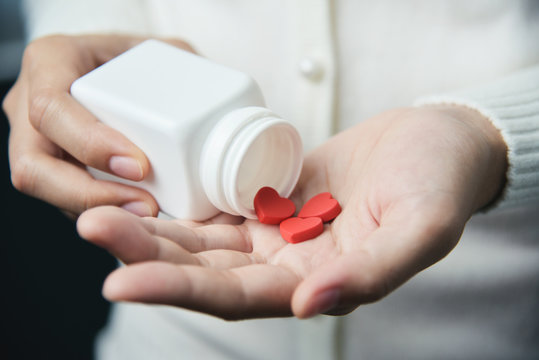 Female hands is holding a white pill bottle and red pills in heart shape.