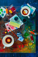 Artist workplace with empty coffee cups, oil and watercolor paint brushes on a colorful palette, sketches and snacks. Flat lay with copy space. Creative occupation concept.