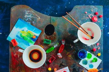 Artist workplace with empty coffee cups, oil and watercolor paint brushes on a colorful palette, sketches and snacks. Flat lay with copy space. Creative occupation concept.