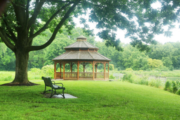 Beautiful summer rainy day nature background. Summer landscape in a city park with a bench on a foreground and wooden gazebo on a background during warm rainy day.