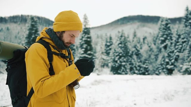 Man with beard, wearing yellow winter clothes using a phone. A hiker goes in the winter mountains with a backpack. Beautiful mountains in winter time