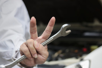 Hands of automotive young mechanic man holding wrench and raising two fingers up it is shows fight with job against car in open hood at the repair garage.