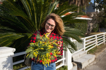 Happy woman with a bouquet of yellow mimosa flowers. Sunny day, the sea coast. The girl laughs, standing near the palm tree.