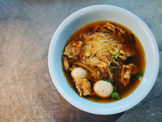 Thai local traditional street food, white small rice noodle in brown clear soup with pig blood, boiled pork and meatball, in blue melamine bowl on stainless steel table, top view from night stall