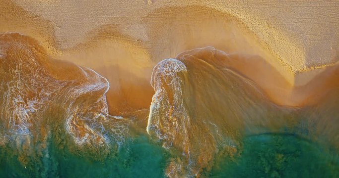Cinematic aerial view of waves breaking on sandy beach at sunset
