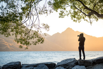 Young male photographer taking photo under the tree at Lake Wakatipu during golden hour sunset in Queenstown, South island, New Zealand, travel and landscape photography concepts