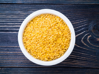 White bowl  with raw bulgur on dark brown wooden background. Top view.