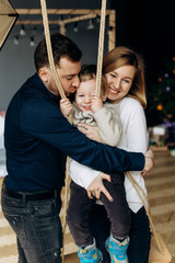 Mom and dad play with charming little son on a swing in the room