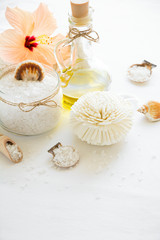 Wellness setting. Sea salt in glass, soap, towel, olive oil and flowers on white textured background. Space for copy. Flat lay