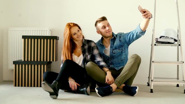 Young couple taking selfie photo with cellphone sitting on floor at their new home
