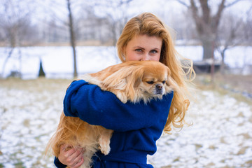 Woman pet owner with blue coat and golden hair walking during winter with her dog. Young lady  with pekingese dog walking on the snowy field. Tender feeling with a dog 