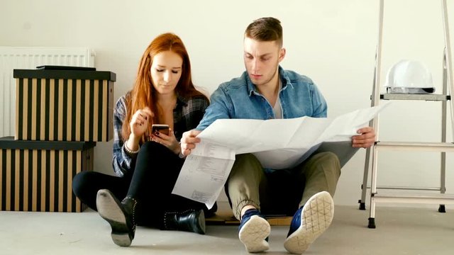 Young couple counting bills with smartphone and blueprints sitting on floor at their new home

