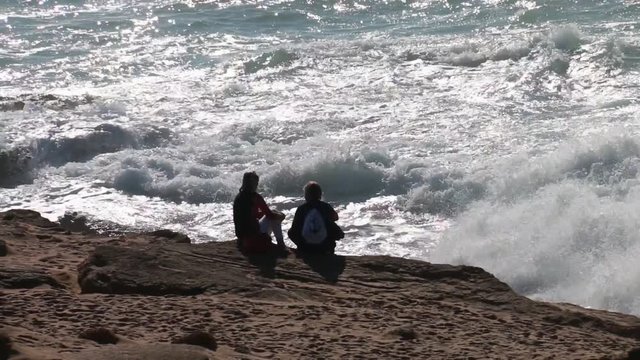 Couple of girls sitting on the edge of a cliff contemplating the strong waves