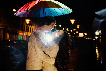 young couple under an umbrella kisses at night on a city street.