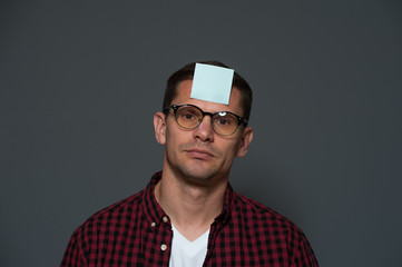 Portrait of sad young man with glasses and shirt with blue sheet glued to his forehead against gray background. Place for text