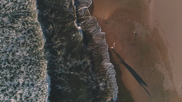 Lonely surfer walk on empty beach, looks for last best wave to catch at warm sunny summer sunset evening. Aerial drone footage of inspiring and beautiful travel place for adventurers