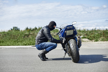Sideways view of motorcyclist in face neck mask sitting on his haunches in the middle of empty road in front of his two wheeled vehicle, studying it before ride. Transport and transportation