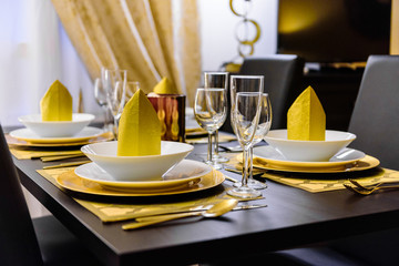 Set table with golden cutlery and napkins, white and golden dishes, different glasses, golden tablecloths and black table. The napkins are on the upper plates as decoration. Golden wall