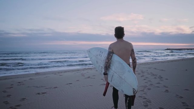 Middle age man in wet suit, authentic surfer walks away into beautiful pink and purple sunset with surf board. Body covered in tattoos, out of office lifestyle choice, freedom and peace concept