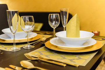 Set table with golden cutlery and napkins, white and golden dishes, different glasses, golden tablecloths and black table. The napkins are on the upper plates as decoration. Golden wall