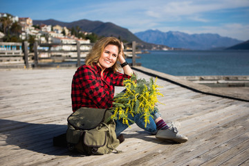 Fototapeta na wymiar Happy girl traveler sits on a wooden pier and holds a bouquet of flowers of mimosa in hands. Next to the woman is a backpack. Sea and mountains on the horizon. Professional lighting.