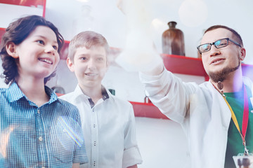 Never get bored. Two amazed boys looking at a fuming laboratory flask held by a male up and coming chemist wearing a laboratory coat and glasses.