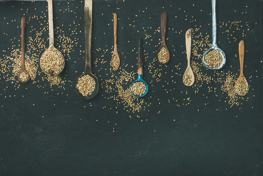 Flat-lay of various old vintage kitchen spoons and green buckwheat grains over black stone background, top view, copy space. Rustic cooking concept