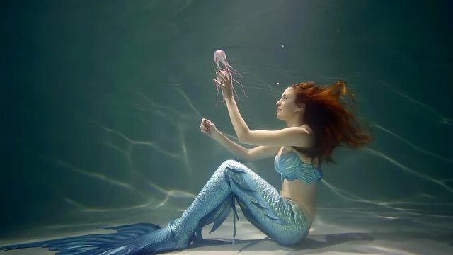 woman with mermaids tail is sitting in an ocean bottom and playing with two pink jellyfishes, touching it