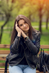 Glamorous young Caucasian woman in black leather jacket sitting in the park on the bench