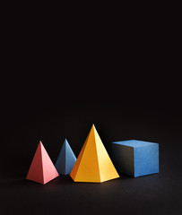 Colorful abstract geometric shape figures still life. Three-dimensional pyramid prism rectangular cube on black background. Yellow blue pink malachite colored objects, copy space