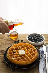 Homemade Belgian waffles served with butter and maple syrup. A hand in motion. Blueberries in the background. 