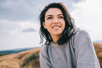 Portrait of happy and funny brunette woman smiling and looking away, against nature meadow and overcast background with windy hair. People, travel and lifestyle concept. Cover mood.