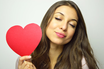 Valentine's Day. Sweet girl in love with closed eyes holding a paper heart and smile at camera on gray background.