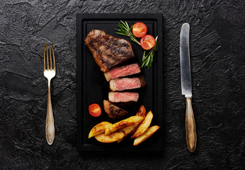 Meat Picanha steak, traditional Brazilian cut with tomatoes, potato wedges and rosemary on black meat cutting board. Steak with fork and knife. Black concrete background.