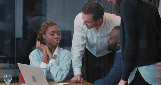 Four attractive colleagues spending evening together in modern office in center of large city. Young friendly people laughing, talking, looking at laptop screen.