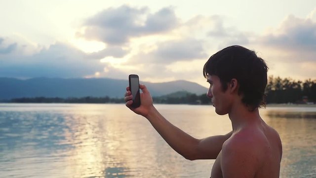 Young handsome man standing on beach taking photo with smart phone of sunset over Koh Samui. Slow motion. 1920x1080