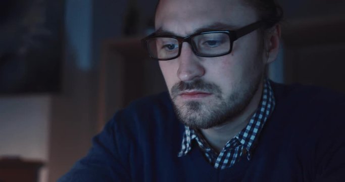 Attractive bearded employee in spectacles sitting in front of computer screen at night. Young man using laptop. Working hard, complicated project. Close up view