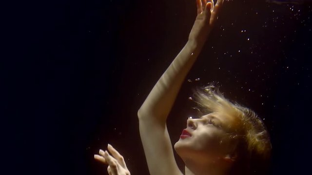 young blonde is swimming under water in fashion bright red dress , pulling hands up to surface