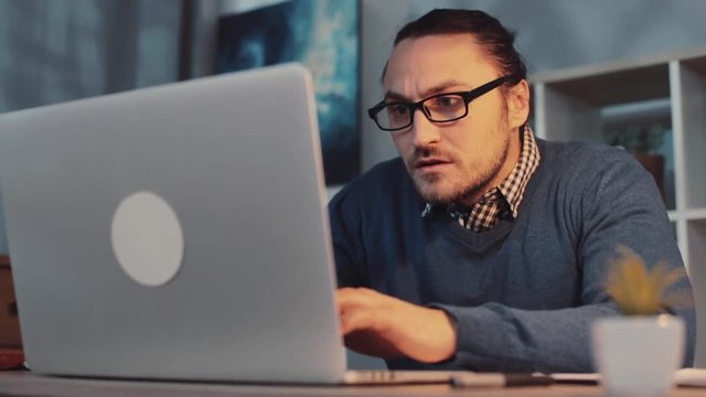 Young man in a stylish look playing computer games on the laptop, he wins, rises his hands, puts off glasses and smiles happily. Gamer, having fun, rest. Virtual reality. Close up view