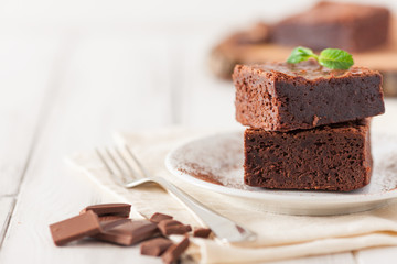 Chocolate brownie square pieces in stack on white plate decorated with mint leaves and cocoa powder...