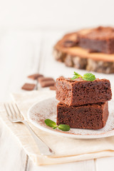 Chocolate brownie square pieces in stack on white plate decorated with mint leaves and cocoa powder on white vintage wooden background. American traditional delicious dessert. Close up photography