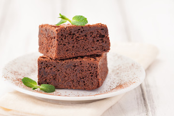 Chocolate brownie square pieces in stack on white plate decorated with mint leaves and cocoa powder on white vintage wooden background. American traditional delicious dessert. Close up photography