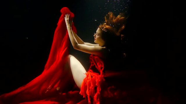 mysterious redhead woman is underwater on black background, playing with train of her bright red dress