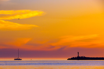 Summer bright orange sunset with a helicopter in the clouds and the silhouette of a sailing boat next to a lighthouse on the shore. Coastal seascape on the Black Sea, Sochi, Russia.
