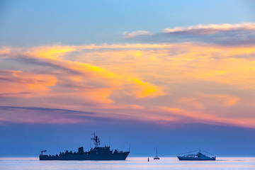 Crossroads on the sea, the silhouette of a warship in front of a boat and a yacht in the light of...