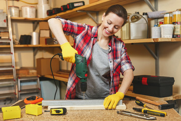 Beautiful caucasian young brown-hair woman in plaid shirt and gray T-shirt working in carpentry workshop at table place, drilling with drill holes in piece of iron and wood while making furniture.
